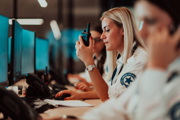 operadora de seguridad femenina sosteniendo radio portátil en la mano mientras trabaja en una sala de control de sistemas de datos oficinas operador técnico trabajando en una estación de trabajo con múltiples pantallas, guardia de seguridad trabajando  - security camera security system surveillance security fotografías e imágenes de stock