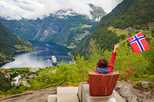 Travel adventure concept. Female tourist enjoying view over Geirangerfjord from Flydalsjuvet viewpoint, holding norwegian flag.