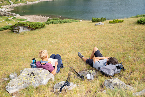 Taking a rest from their hiking, a young couple lay down for a rest on the hillside, resting against some rocks to enjoy the view of the lake