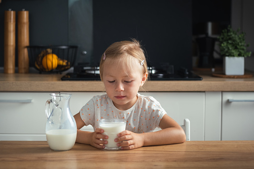 Sad little caucasian girl refuses to drink milk in the stylish kitchen. Health benefits of milk for little kids concept