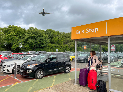 London, England - May 2022: Person with suitcases waiting at a bus stop in the long stay parking area. A plane is flying ovehead.