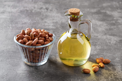 Peanut oil in a glass jug and raw peeled groundnut in a glass bowl over gray background. Arachis hypogaea as edible seeds and oil crop. Monounsaturated cooking oil. Vegetarian snack. Top view.