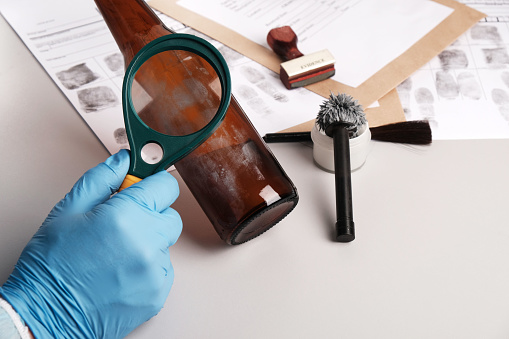 forensic expert using a magnifying glass examines fingerprints on evidence -  glass bottle, forensic fingerprint analysis in  police forensic laboratory