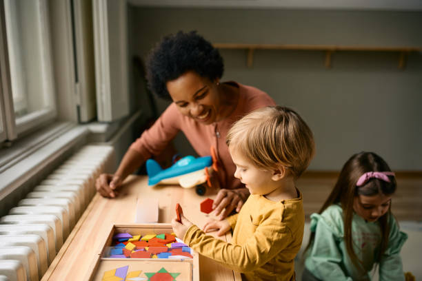 Happy boy playing with colorful puzzle shapes with his kindergarten teacher. Happy kid and his African American teacher using wooden puzzle shapes while playing at kindergarten. nanny stock pictures, royalty-free photos & images