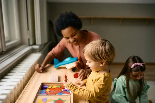 Happy kid and his African American teacher using wooden puzzle shapes while playing at kindergarten.
