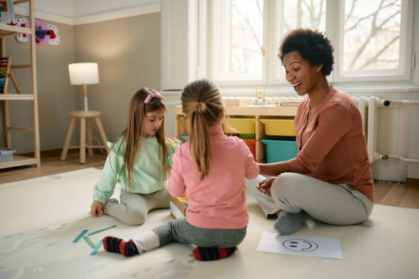 Happy black teacher and small girls having fun while playing at preschool. Happy African American preschool teacher and little girls having fun while sitting on the floor and playing. nanny stock pictures, royalty-free photos & images