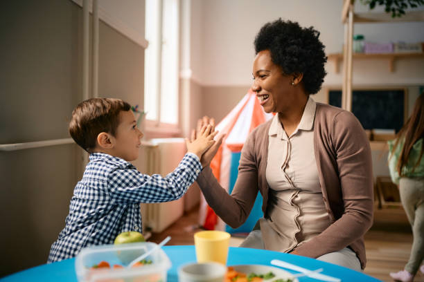 Happy black teacher and small boy giving high five during lunch at preschool. Happy little boy giving high-five to his African American teacher after eating lunch at kindergarten. preschool teacher stock pictures, royalty-free photos & images