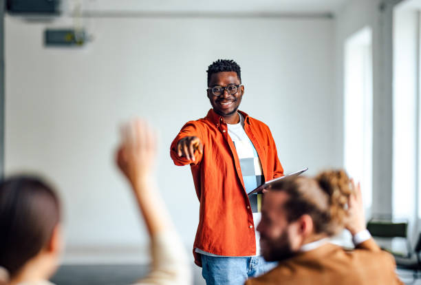 Cheerful Male Presenter Interacting With the Audience Handsome cheerful man in a orange shirt standing in front of an audience holding a tablet and using hand gestures to interact with the audience. instructor stock pictures, royalty-free photos & images