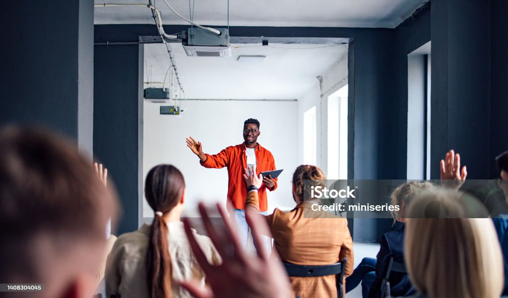 Cheerful Male Presenter Interacting With the Audience Handsome cheerful man in a orange shirt standing in front of an audience holding a tablet and using hand gestures to interact with the audience. Public Speaker Stock Photo