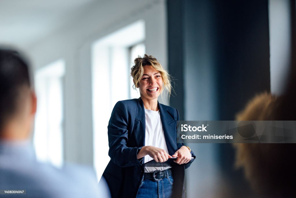 Cheerful Female Presenter Interacting With the Audience Beautiful cheerful woman in a dark blue jacket standing in front of a crowd looking down at a tablet. Women Stock Photo