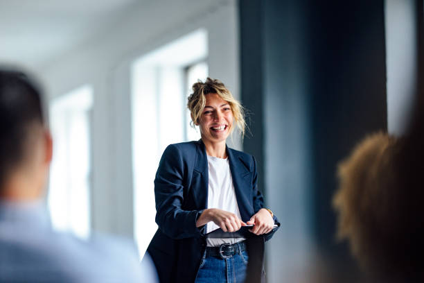 Cheerful Female Presenter Interacting With the Audience Beautiful cheerful woman in a dark blue jacket standing in front of a crowd looking down at a tablet. presenter stock pictures, royalty-free photos & images