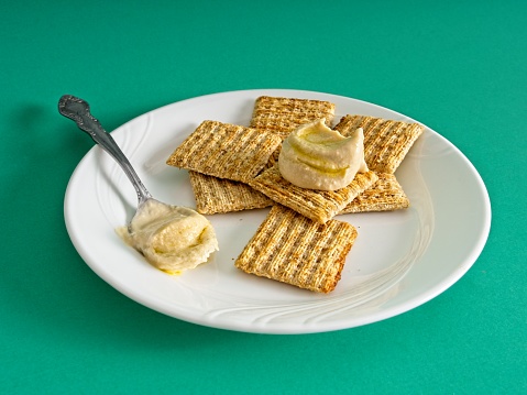 Whole wheat crackers and freshly prepared hummus with olive oil on a white plate. A simple vegan snack of whole grains and blended chickpeas and olive oil. With serving spoon on a green background with copy space.