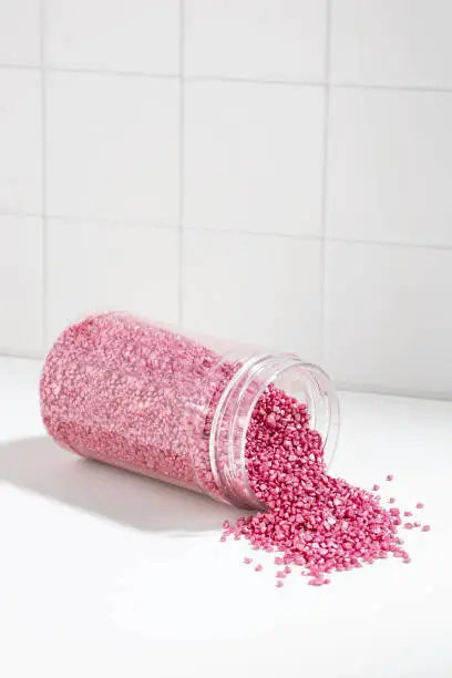 Package of pink shiny crystal salt on the background of the bathroom wall. Jar with scattered shimmering pink sea salt. Idea of relaxation, aromatherapy and self care. The effect of sea water on hair.