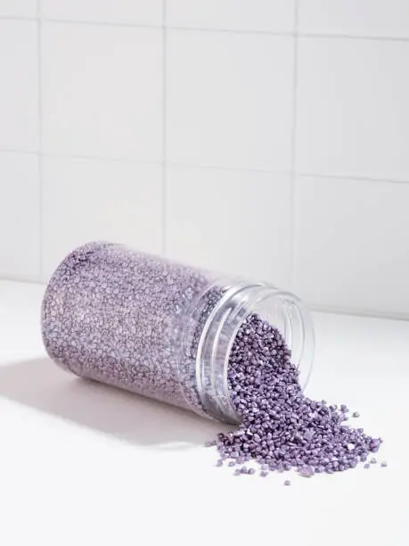 Package of purple glitter crystal salt on a background of the bathroom wall. Jar with scattered shimmering purple sea salt. Idea of relaxation, aromatherapy and self care. Effect of sea water on hair.