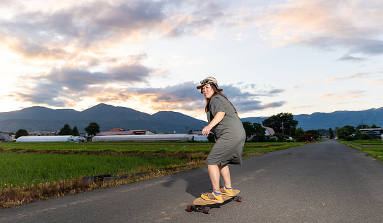 A mid age Japanese woman skateboards along a quiet country road.