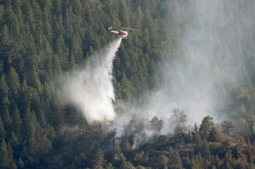 Flying low over a large group of firefighters in yellow, a fire fighting helicopter drops water on the Snowcreek fire near Highway 285 on July 12, 2022 in a pine forest in the Rocky Mountain foothills of Morrison, Colorado.