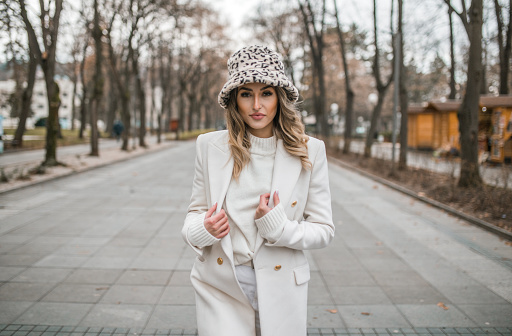 A beautiful young woman dressed in a white wardrobe walking through the park during a winter autumn day, before the New Year holidays.