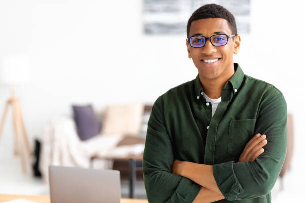 smiling successful businessman with eyeglasses stands with crossed arms smiling and looking at the camera. young male confident entrepreneur or small business owner in office, copy space - business looking at camera office new imagens e fotografias de stock