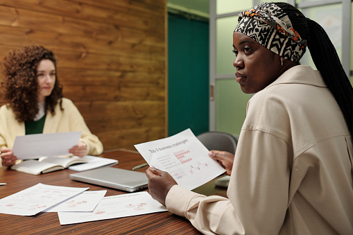 Young black woman with financial document looking at one of colleagues while sitting by workplace during discussion at working meeting