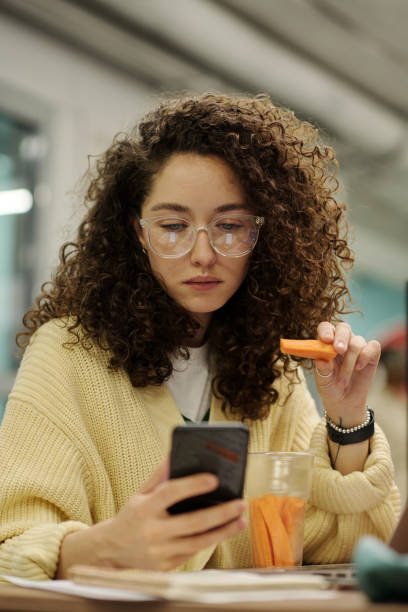 Young serious businesswoman with smartphone eating carrots for lunch and texting stock photo