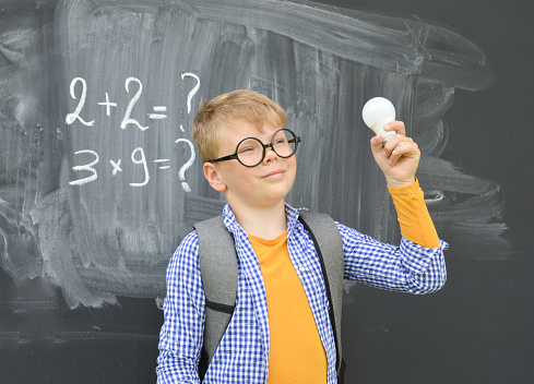 Portrait of a Caucasian boy on the background of a black school blackboard, who holds a light bulb in his hands. Ideas, inspiration, learning concept.