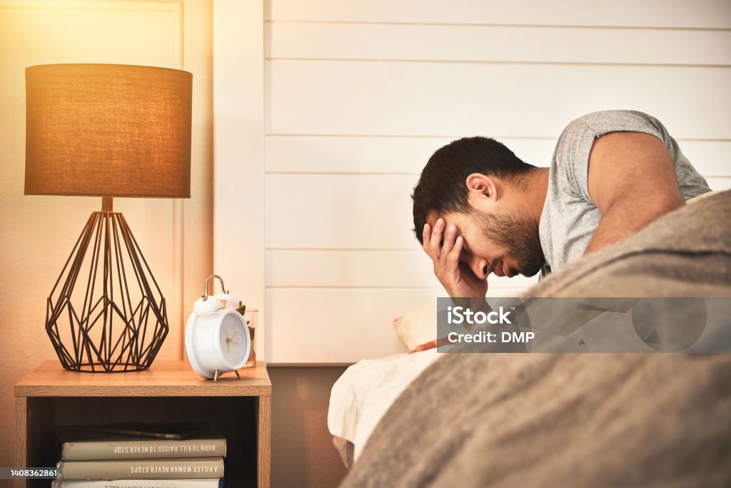 Young displeased, tired man with insomnia unable to sleep and suffering from burnout and fatigue struggling to get up out of bed in the morning. Mentally exhausted male awake in bed feeling stressed Sleeping Stock Photo