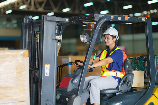 Asian beautiful woman driver drive fork lift car in industry with smile, ability of girl and diversity of career wearing helmet in reflective vest working on vehicle