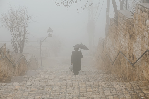 An elderly man in a hat and coat walks up the stairs in the fog under a black umbrella. Lonely sad man in scarf with umbrella walking under rain. Back view of an old man. Sadness, longing, loneliness.