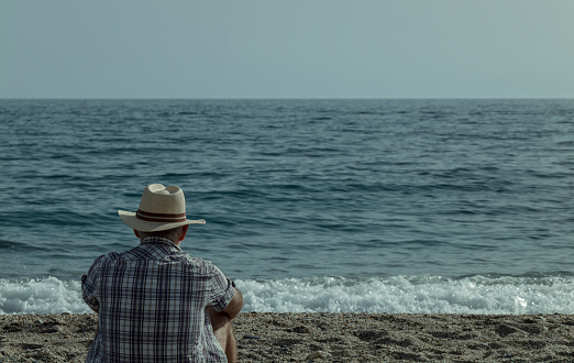 Rear view of adult man in hat with shirt sitting on beach