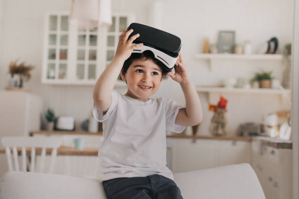 istock Little cheerful boy smiling wide taking off vr glasses, amazed after using virtual reality googles sitting on sofa over blurry kitchen at home. Playfull Spanish boy at home using vr lenses for games. 1408368162