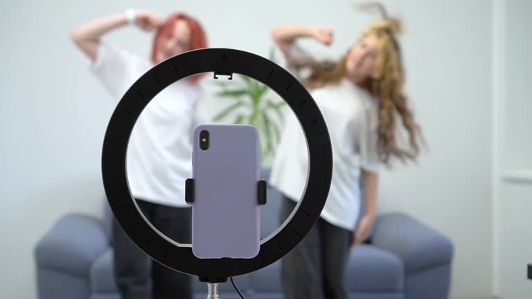 Two white teenage girls dance and film a viral video for their social media blog. Focus on the phone. The smartphone is mounted on a tripod with a lamp.