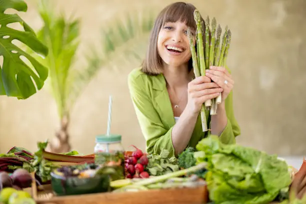 Portrait of a young cheerful woman holds a bunch of asparagus while sitting by the table full of fresh vegetables, fruits and greens. Healthy eating and lifestyle concept