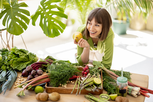 Portrait of a young cheerful woman stands by the table full of fresh vegetables, fruits and greens with plants on background indoors. Healthy eating and lifestyle concept