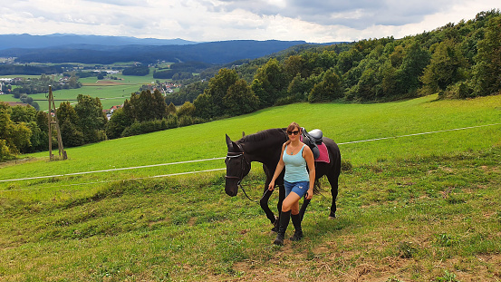 PORTRAIT: Cheerful young Caucasian woman leads her horse by the rein up a grassy hill. Female horseback rider leads her chestnut stallion from ranch enclosure. Girl exploring countryside with horses