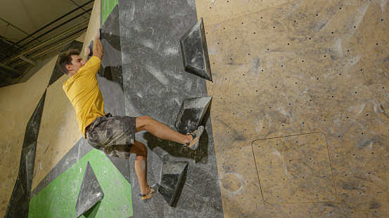 COPY SPACE: Flexible male climber skilfully navigates a challenging indoor climbing route. Athletic young man holds on to black volume holds while bouldering at a colorful indoor training facility.