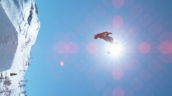 VERTICAL, LENS FLARE: Fearless male tourist snowboarding in fun park does a big backflip on a perfect winter day. Breathtaking shot of unrecognizable man snowboarding and doing a spectacular stunt.