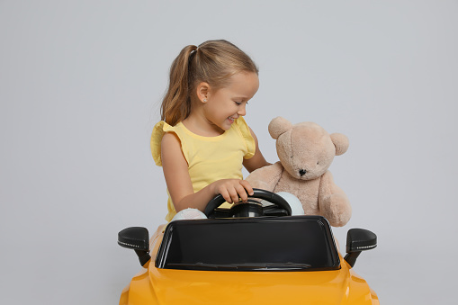 Cute little girl with toy bear driving children's car on grey background