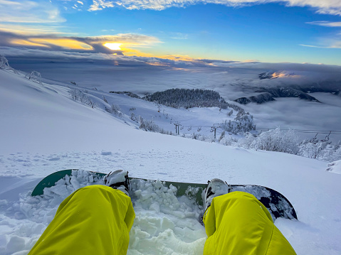 POV: Looking at the golden sunset while sitting in the fresh powder snow during a snowboarding trip in the Julian Alps. Stopping during an offpiste snowboarding session to watch the scenic sunrise.