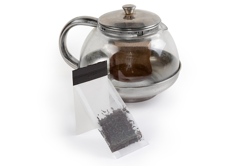 Tea bag of dry black tea leaves made with food plastic mesh rectangular shape with attached paper label on a blurred background of the glass teapot on a white surface