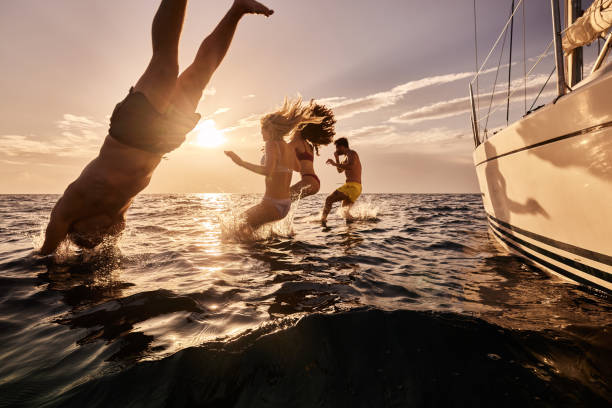 des gens insouciants qui sautent dans la mer depuis le bateau au coucher du soleil. - sailboat sunset sailing nautical vessel photos et images de collection