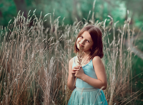 A 10-year-old girl stands near tall dry grass and holds a bouquet in her hands. The girl is holding a bouquet. The girl is wearing a brown t-shirt and a blue skirt.