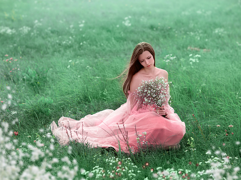A young girl walks in a field of white flowers. The girl is holding a bouquet in her hands. The girl is wearing a pink long dress.