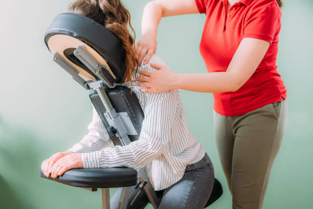Female employee sitting on a portable massage chair in business office. Therapist massaging her shoulders, releasing muscle tension. stock photo