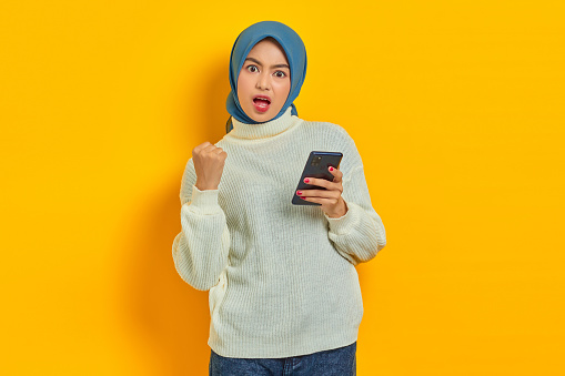 Shocked young asian woman in white sweater and hijab using on mobile phone,  do winner gesture clench fist say yes isolated over yellow background
