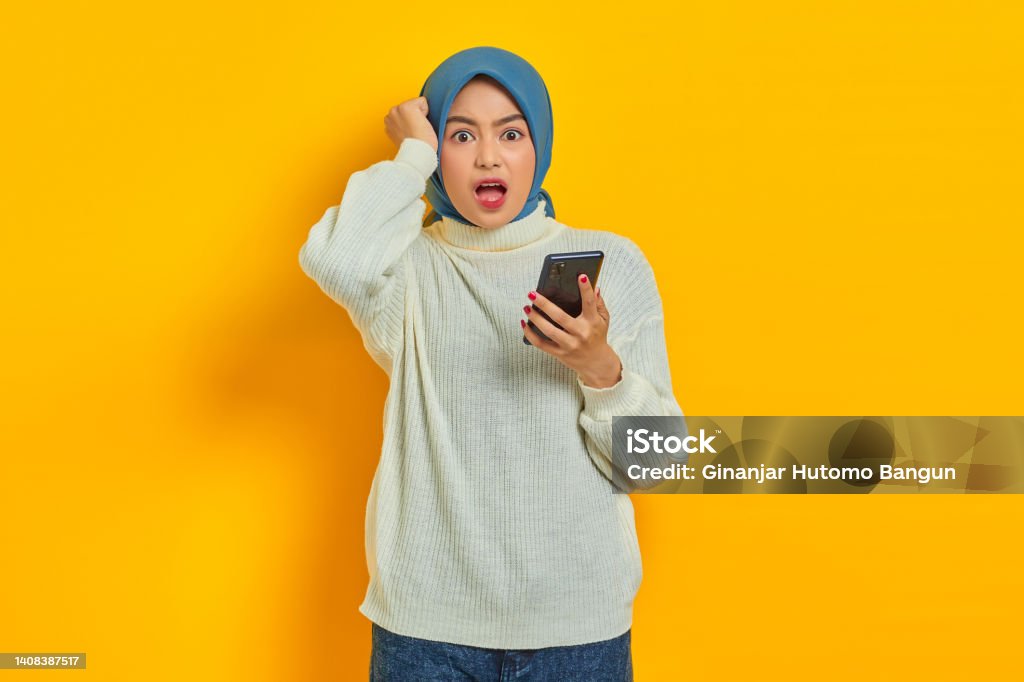 Shocked young asian woman in white sweater and hijab using on mobile phone and holding head with hand isolated over yellow background 20-24 Years Stock Photo