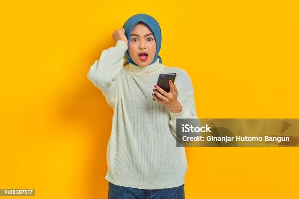 Shocked Young Asian Woman In White Sweater And Hijab Using On Mobile Phone And Holding Head With Hand Isolated Over Yellow Background Stock Photo - Download Image Now