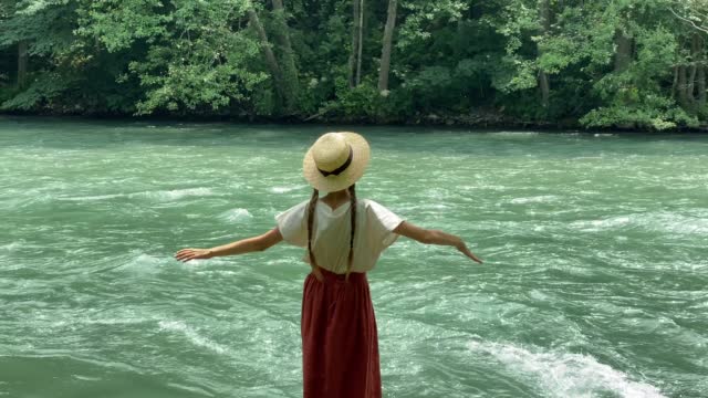 Young beautiful woman in long skirt and straw boater hat standing on stones near powerful rippling mountain river and dancing. Summer travel concept, romantic vintage girl, old retro style clothes