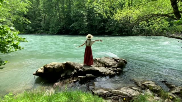 Young beautiful woman in long skirt and straw boater hat standing on stones near powerful rippling mountain river and dancing. Summer travel concept, romantic vintage girl, old retro style clothes