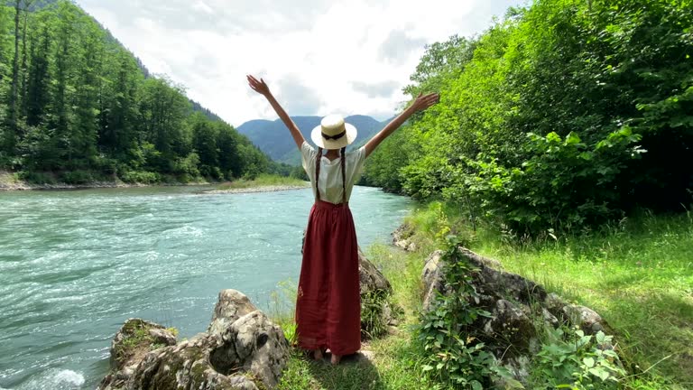 Young beautiful woman in long skirt and straw boater hat goes to stone coast near powerful rippling mountain river and raises hands up. Summer travel concept, romantic vintage girl, old retro style