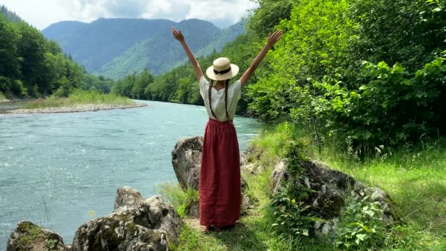 Young beautiful woman in long skirt and straw boater hat goes to stone coast near powerful rippling mountain river and raises hands up. Summer travel concept, romantic vintage girl, old retro style